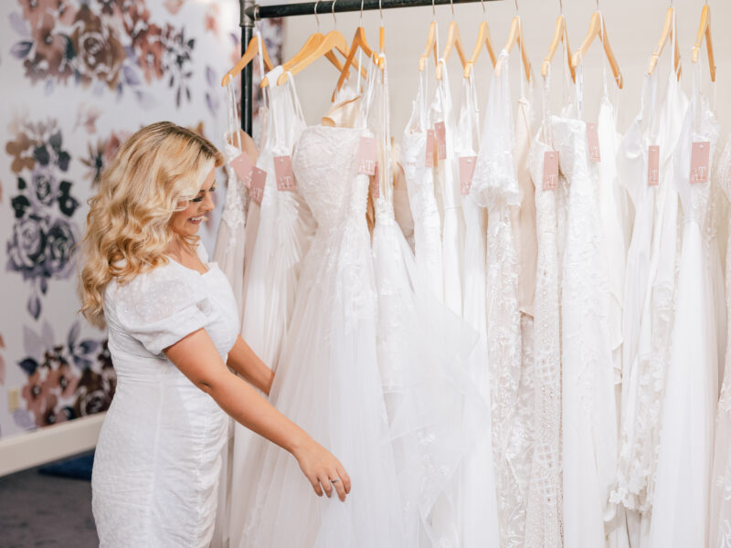 smiling bridal stylist inspecting white bridal gowns on rack