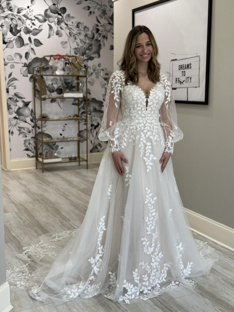 A smiling bride wearing a flowy wedding dress with lace sleeves and leaf detailing