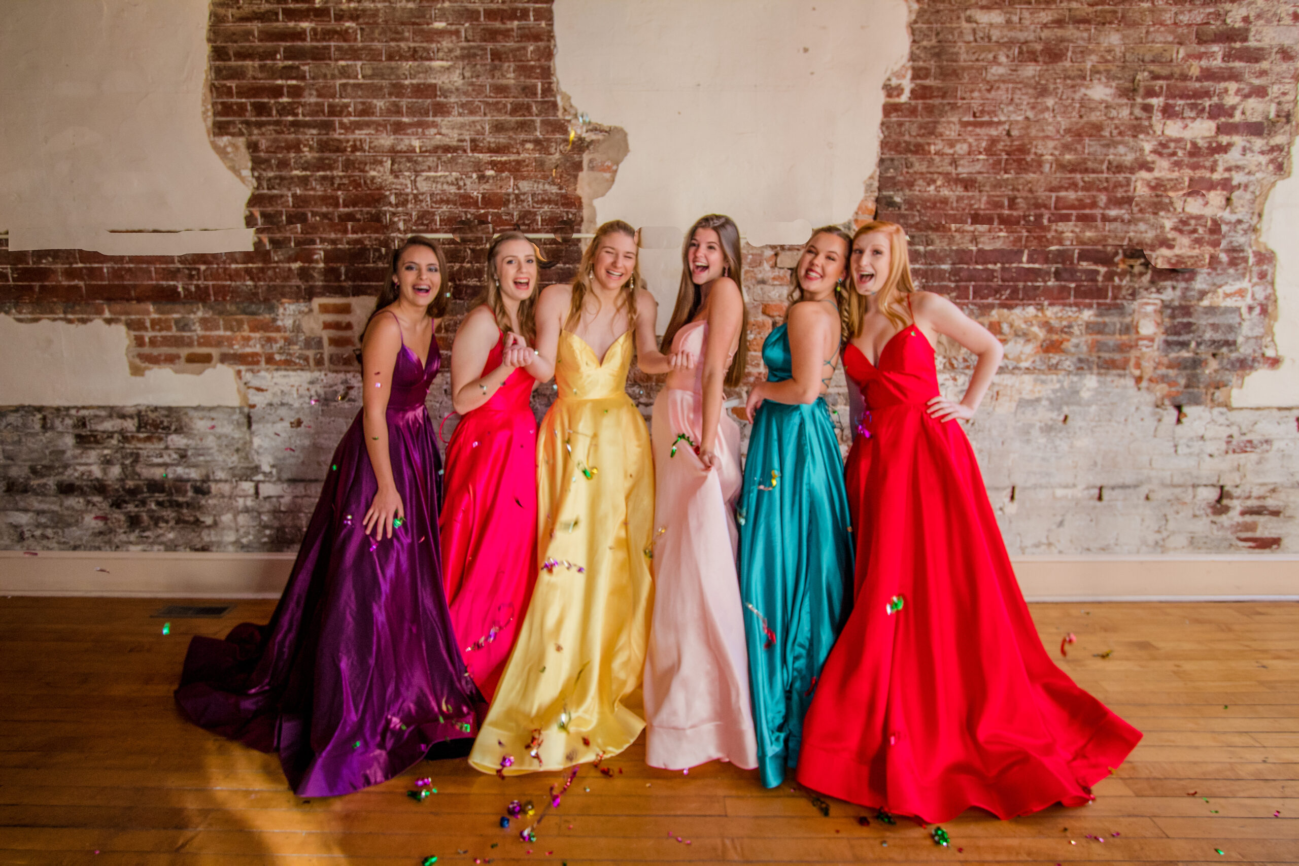 6 girls standing in multicolored prom dresses throwing confetti and laughing in a brick building