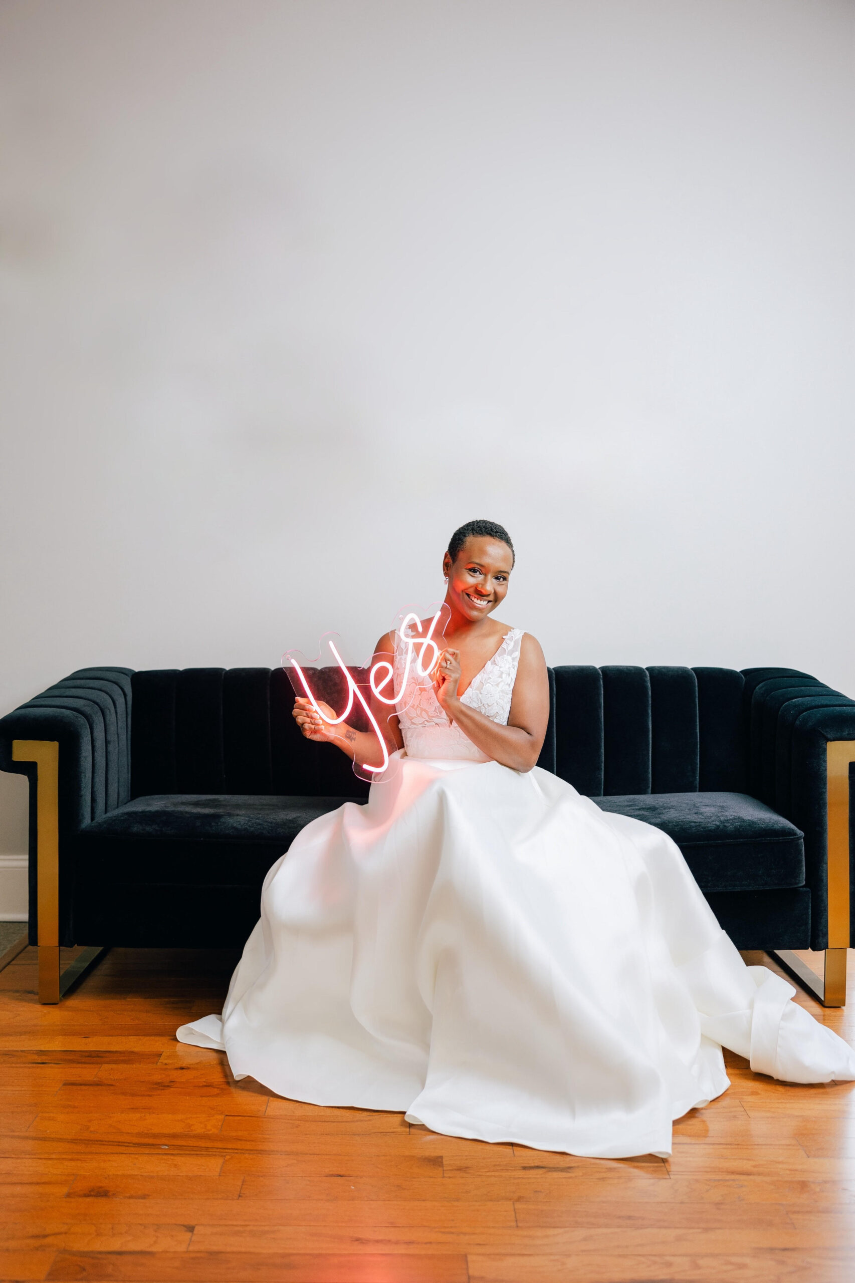 bride sitting on black couch smiling in her wedding gown holding a neon sign that says "Yes"
