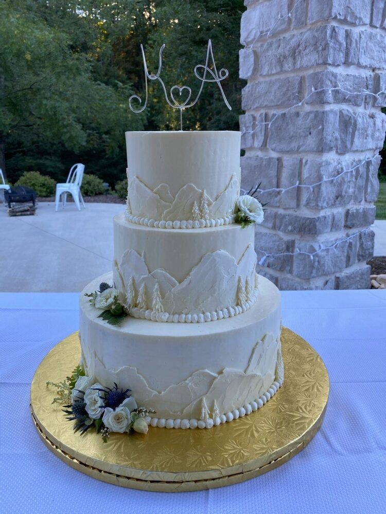 A three-tiered wedding cake with white icing and edible mountain decorations going around each tier of cake.