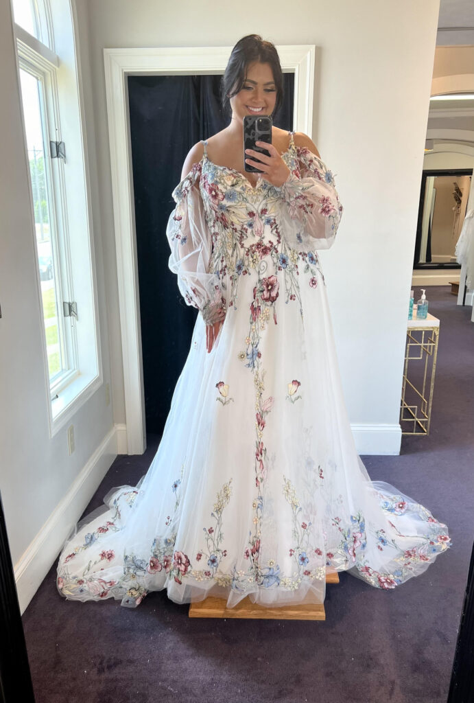 A bride taking a selfie in the mirror at Amanda's Touch wearing a white wedding dress with colorful floral details