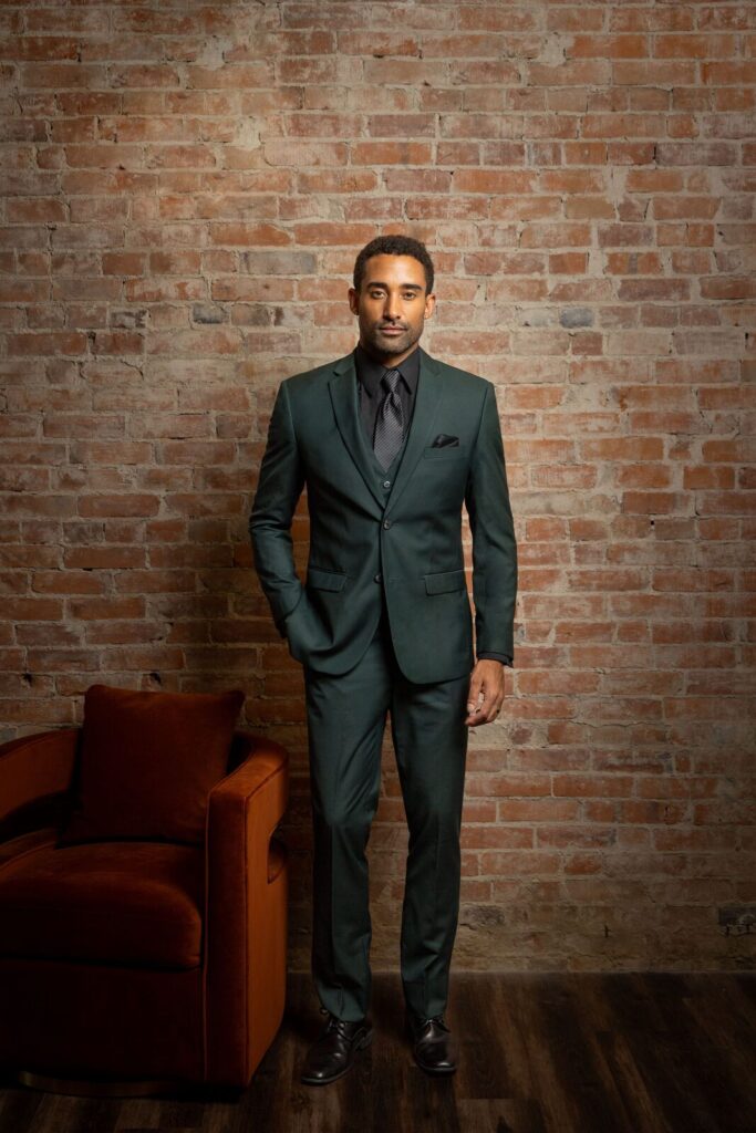 A man in a dark green tuxedo a color known as "hunter green" standing in front of a brick wall