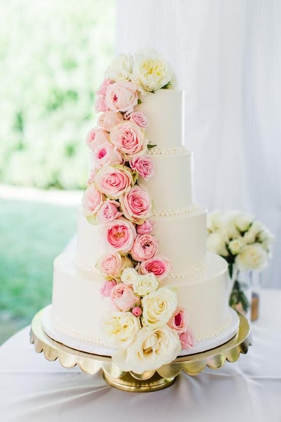 A four-tiered wedding cake with elegant white icing and a dozen rose decorations lining the side of the cake, on a gold platter.