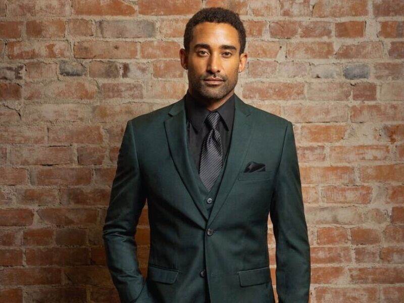 A man in a dark green tuxedo a color known as "hunter green" standing in front of a brick wall