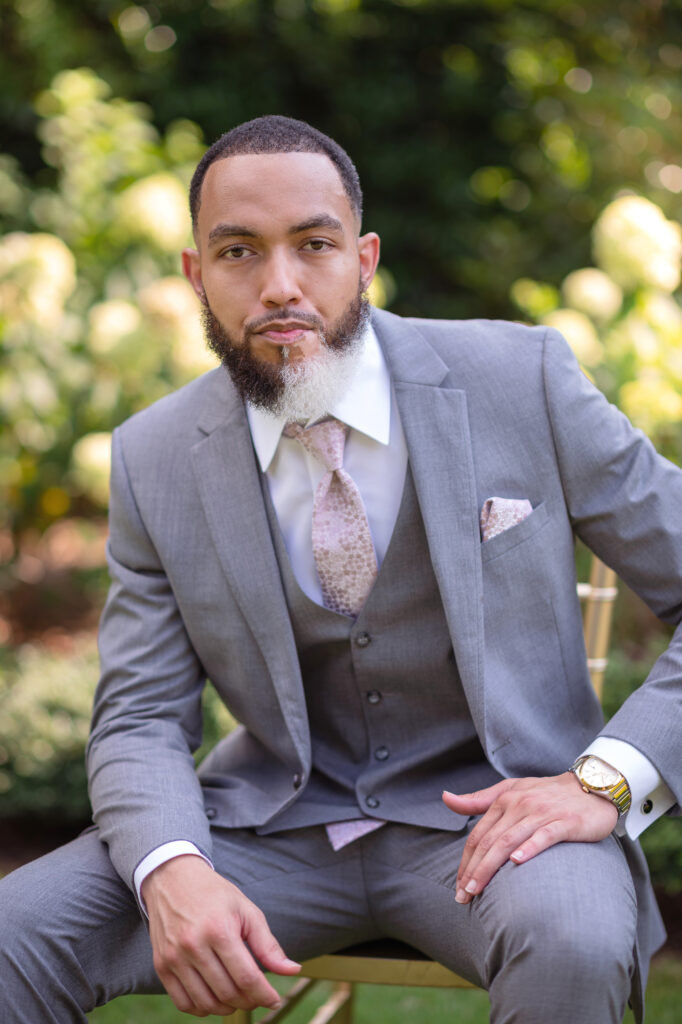 A man seated in a dapper gray tuxedo suit and a pink tie with a serious expression on his face