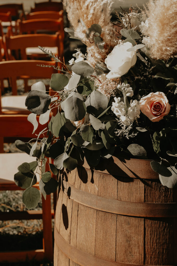 Rustic wedding decoration on a barrel with flowers and greenery under sunlight.