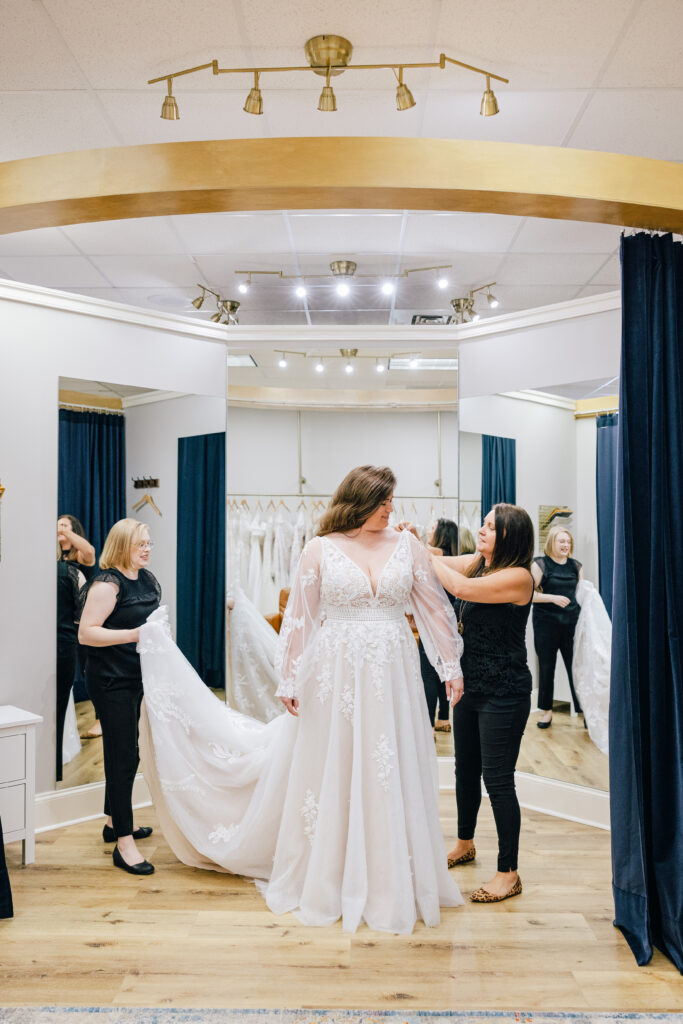 Bride in lacy gown fitted in a bridal salon with assistants adjusting the dress.