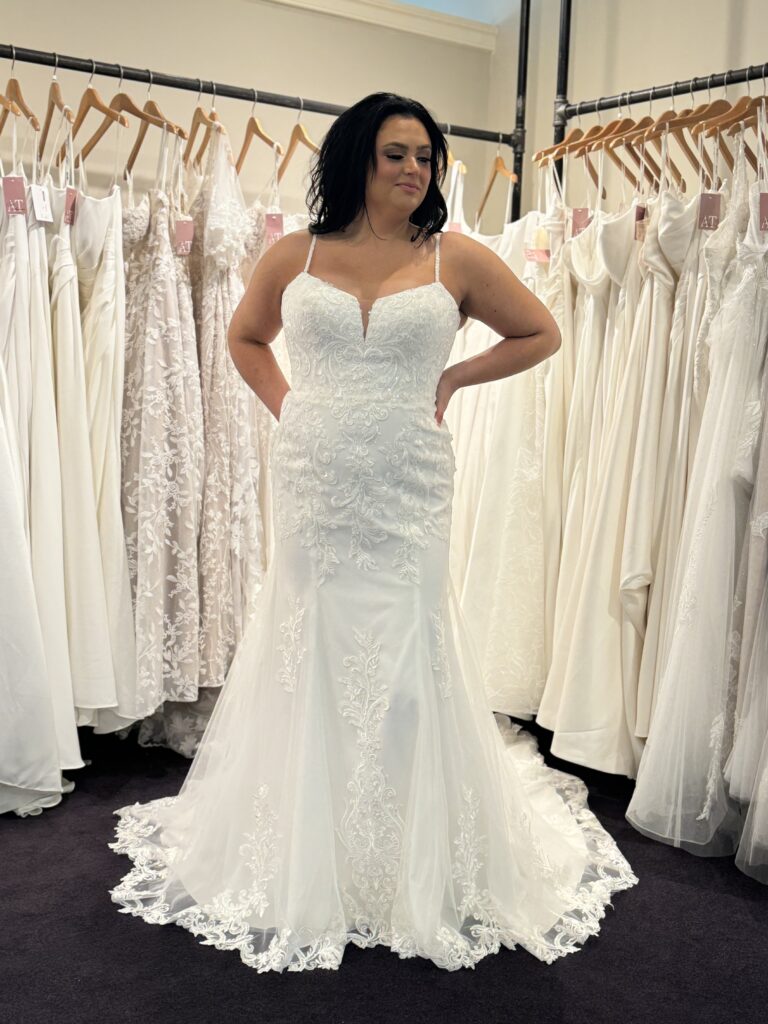 Confident bride poses in a fitted lace wedding dress in a well-lit bridal store.