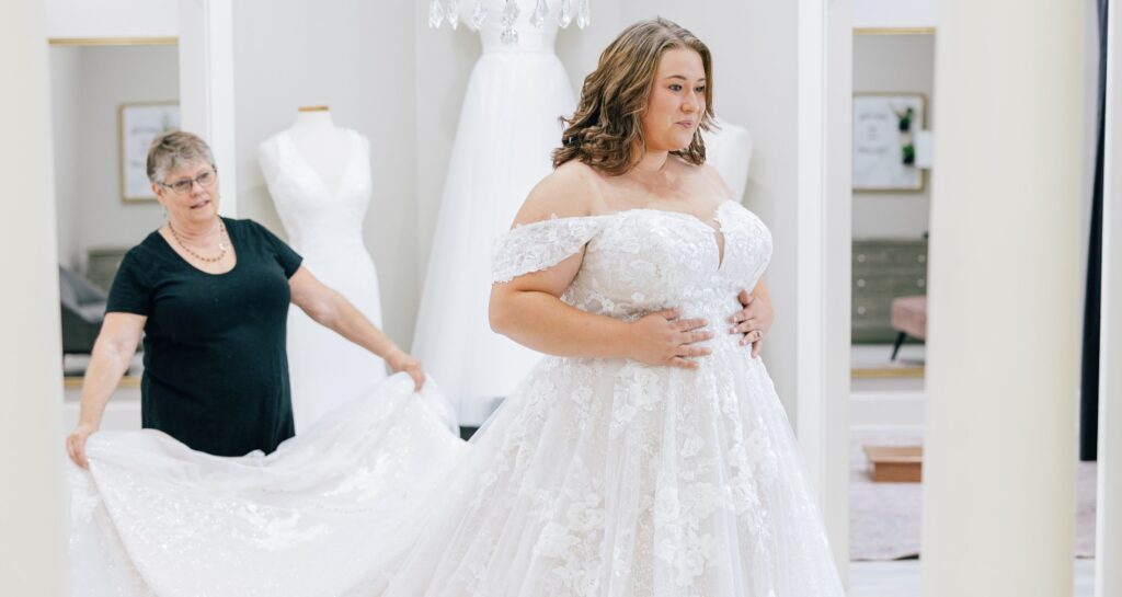 Bride in a detailed lace gown smiles as she is assisted with her dress in a boutique.