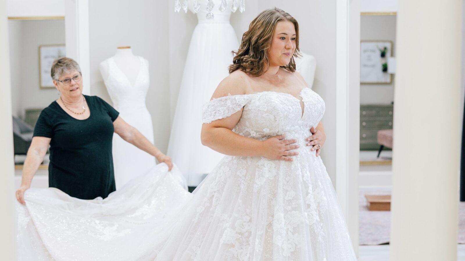 Bride in a detailed lace gown smiles as she is assisted with her dress in a boutique.