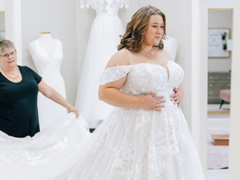 Bride in a detailed lace gown smiles as she is assisted with her dress in a boutique.