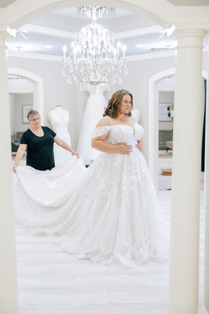 Bride in elaborate white gown admires herself in boutique mirror, assistant helps with train.