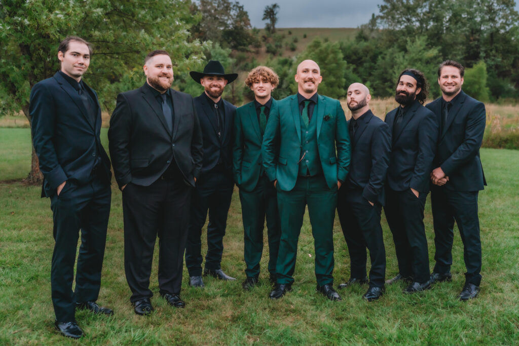 Group of groomsmen, two in emerald-green suits, others in black, posing outdoors with a relaxed, rustic background.