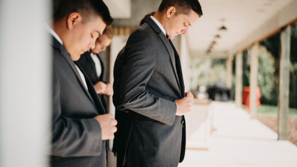 Groomsmen in gray suits adjusting their jackets, standing under a covered walkway with soft natural lighting.