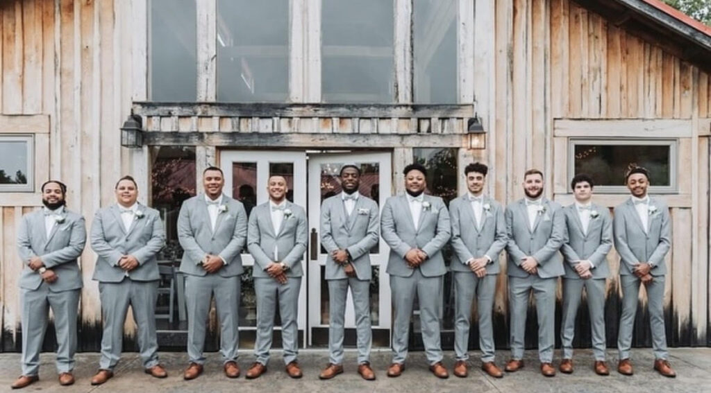 A lineup of groomsmen wearing light gray suits and brown shoes standing in front of a rustic wooden building.