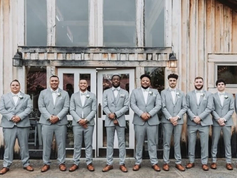 A lineup of groomsmen wearing light gray suits and brown shoes standing in front of a rustic wooden building.