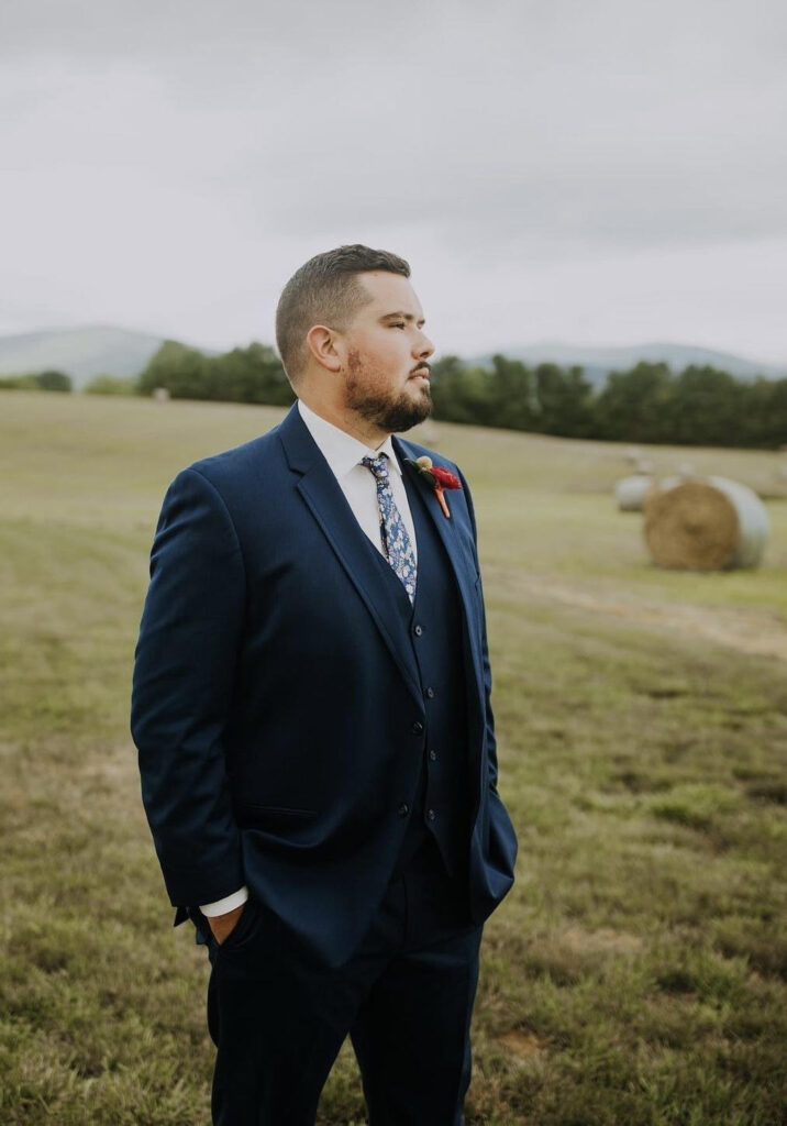 Groom in a navy-blue suit with a floral tie and boutonniere, standing outdoors in a grassy field with a distant view.