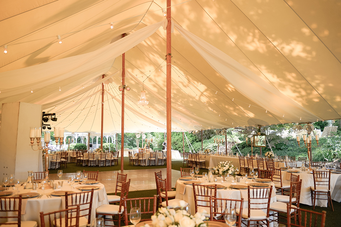 Winery wedding reception tables with candles and floral arrangements under a tent with lights.