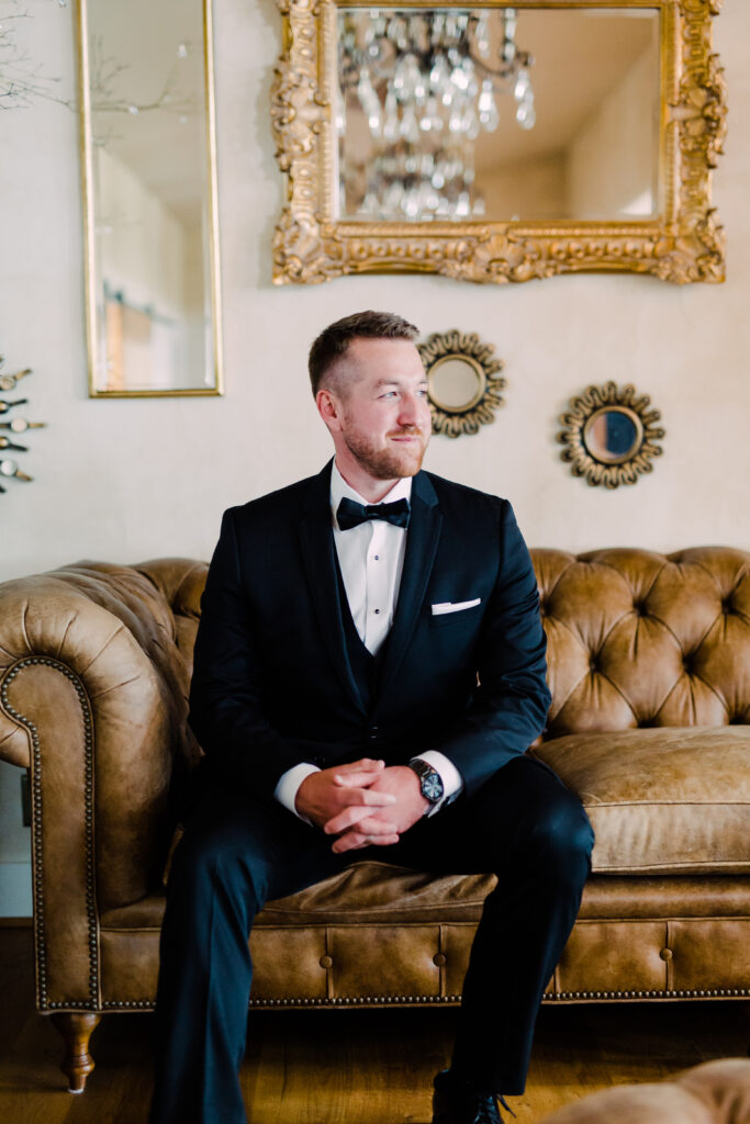 Groom seated on a vintage leather couch in a black tuxedo and bow tie, with decorative mirrors on the wall behind.