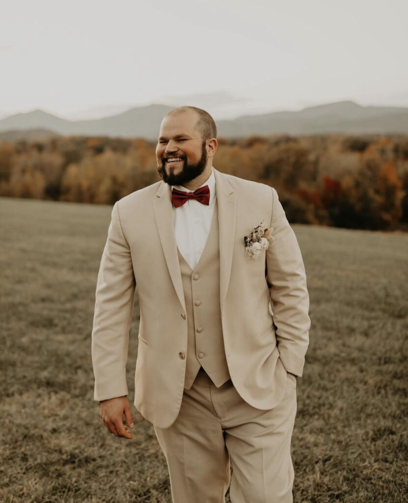 Groom in a beige suit with a red bow tie and boutonniere, smiling against a scenic fall landscape background.