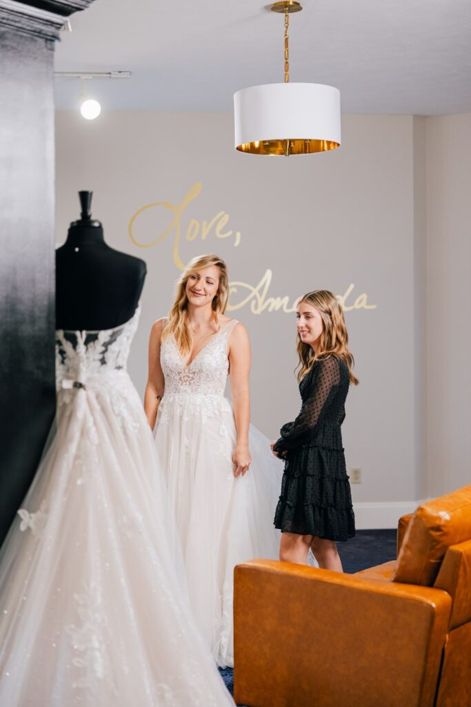 A bride-to-be modeling an elegant lace wedding dress in the boutique, alongside a stylist offering advice and support.