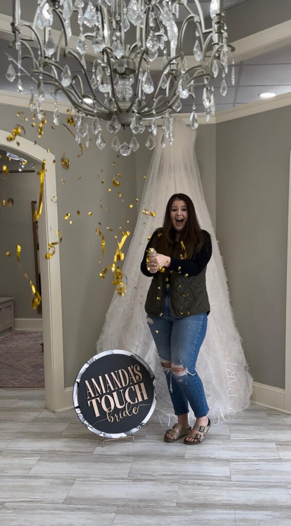 A happy bride popping golden confetti under a sparkling chandelier at Amanda's Touch bridal boutique, standing by a bridal gown display.