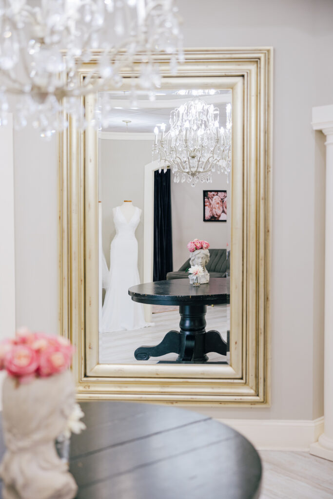 A golden-framed mirror reflecting a stunning wedding dress on a mannequin, accented by elegant chandeliers and décor.