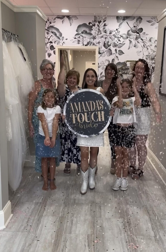 A bride surrounded by her family and friends celebrating at Amanda's Touch bridal shop, holding a custom "Amanda's Touch Bride" sign.