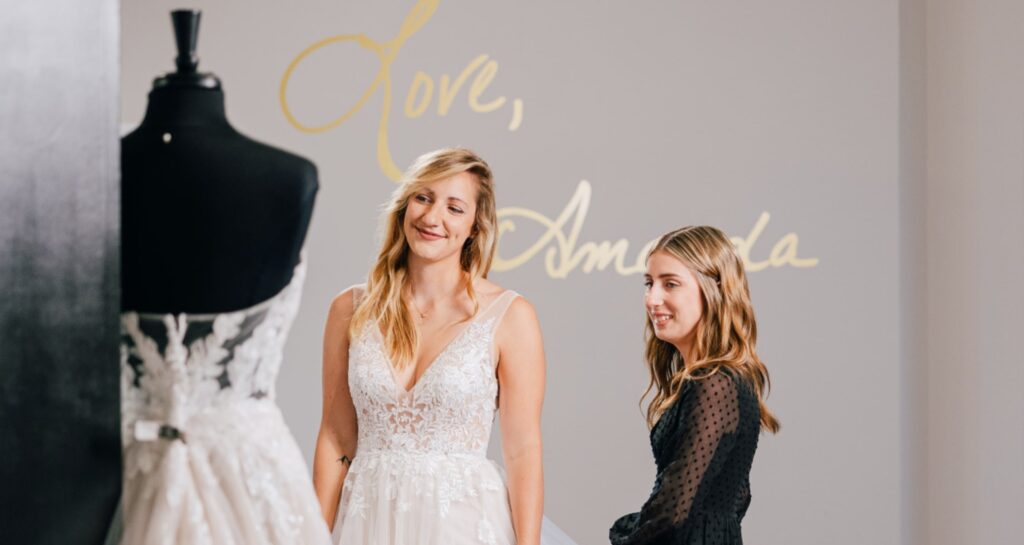 A bride-to-be smiles while trying on a lace wedding gown at Amanda's Touch bridal boutique with assistance from a stylist.