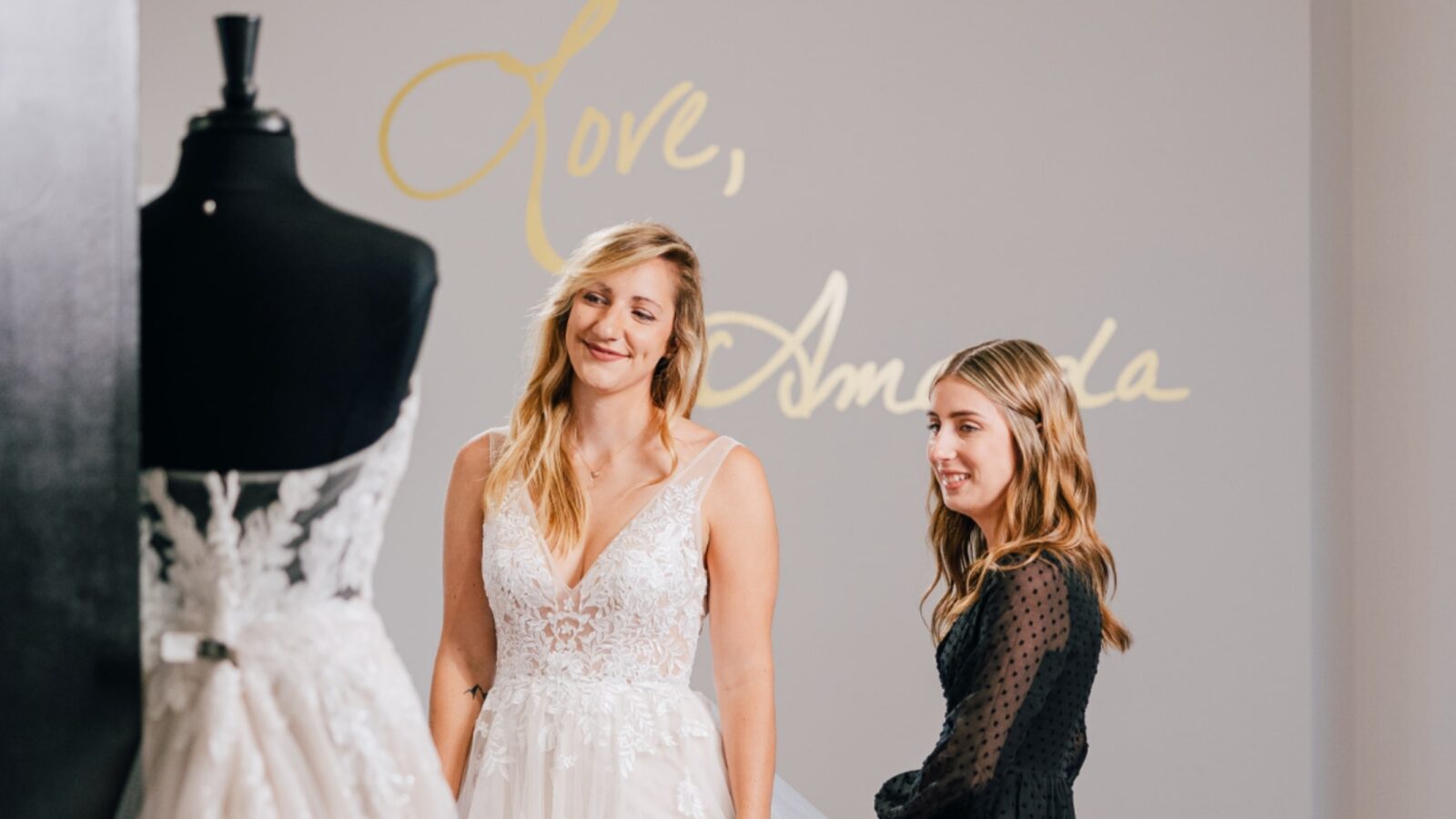 A bride-to-be smiles while trying on a lace wedding gown at Amanda's Touch bridal boutique with assistance from a stylist.