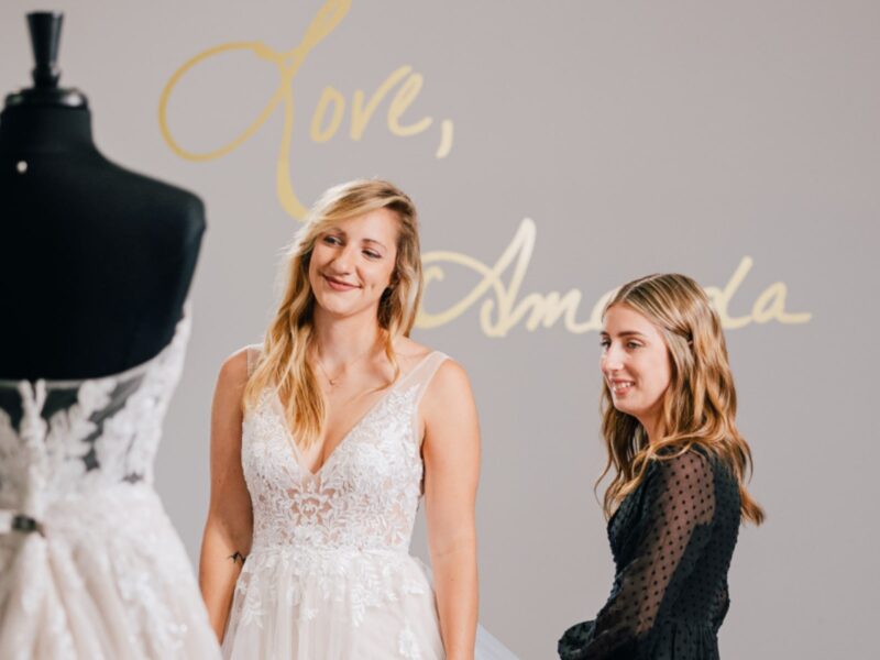 A bride-to-be smiles while trying on a lace wedding gown at Amanda's Touch bridal boutique with assistance from a stylist.