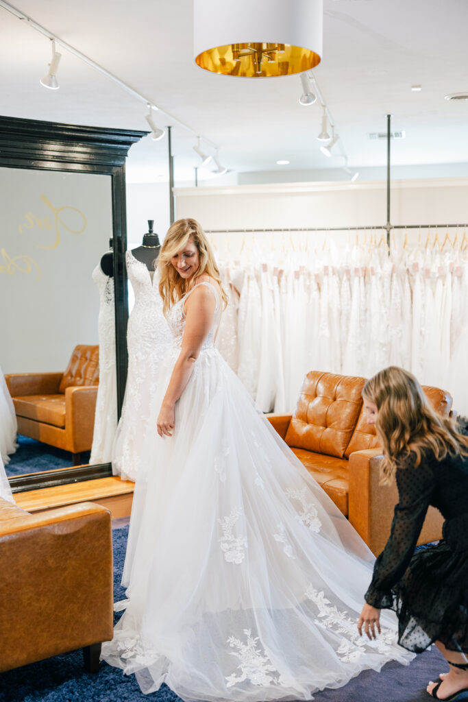 A bride standing in a long, flowing lace wedding gown as a stylist adjusts the train in a bright, luxurious bridal boutique.