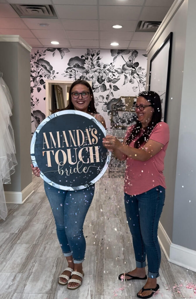 A bride surrounded by her family and friends celebrating at Amanda's Touch bridal shop, holding a custom "Amanda's Touch Bride" sign.