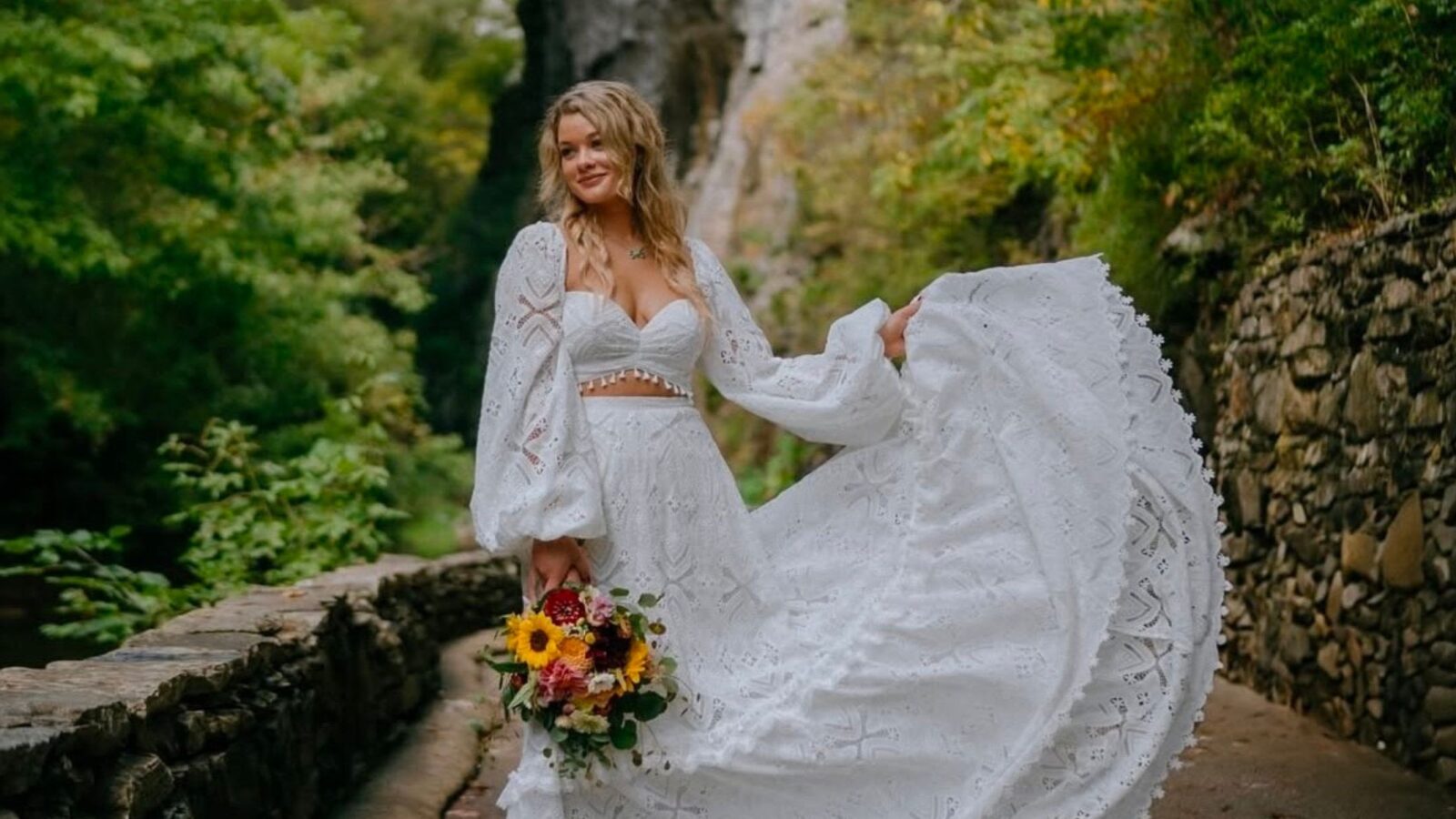 Bride wearing a stunning white boho lace dress with balloon sleeves, holding a colorful bouquet, smiling near a scenic stone path.
