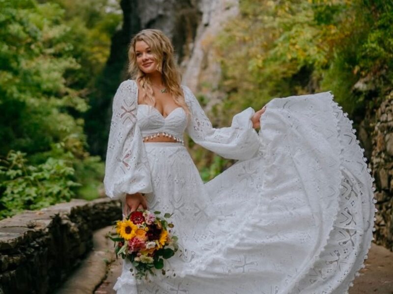 Bride wearing a stunning white boho lace dress with balloon sleeves, holding a colorful bouquet, smiling near a scenic stone path.