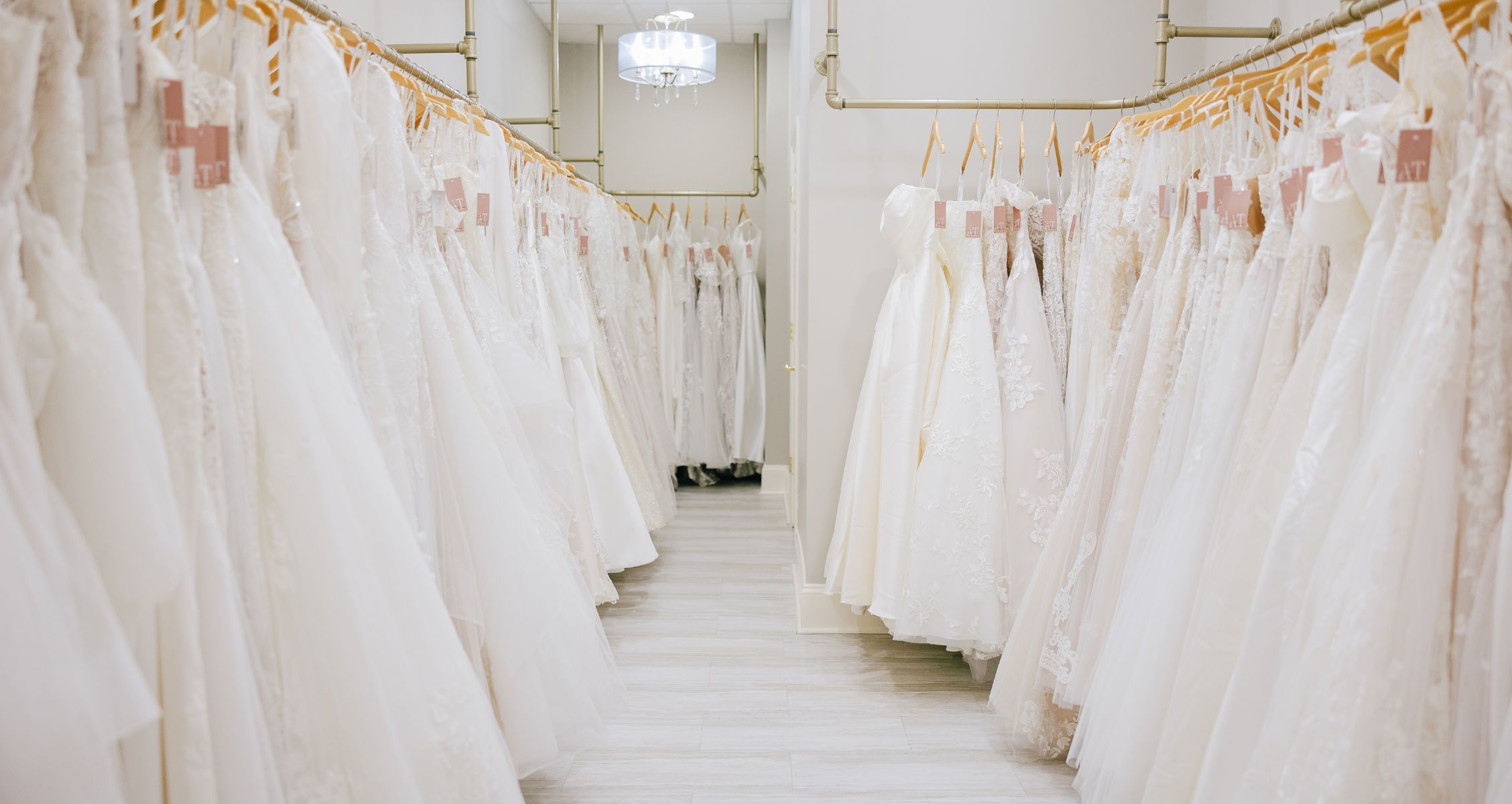 A well-lit bridal boutique aisle showcasing rows of elegant wedding dresses on wooden hangers, organized neatly with a chandelier overhead.