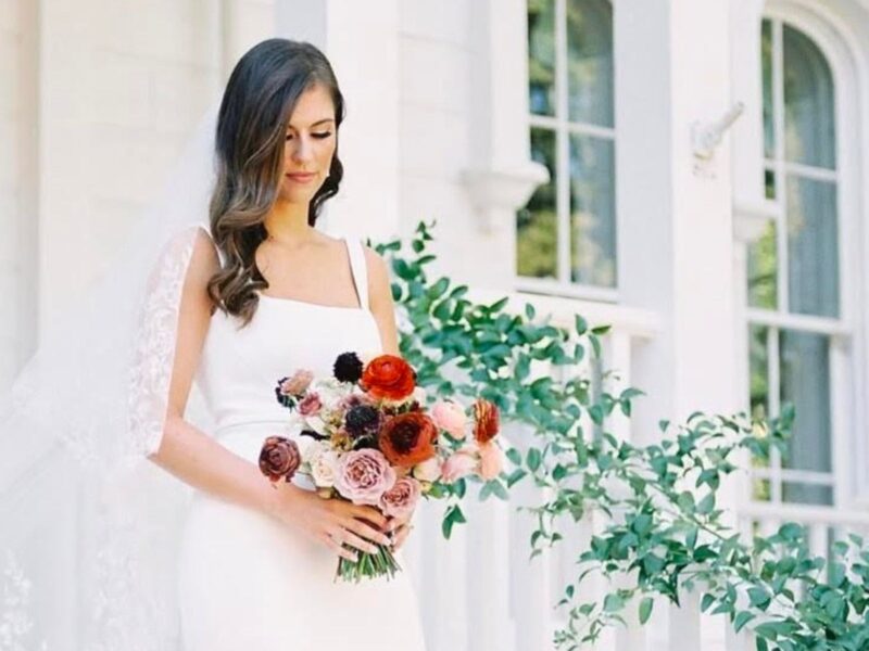 A bride stands on the steps of a white historic building, wearing a sleek white gown and holding a bridal bouquet featuring rich red, blush, and peach blooms.