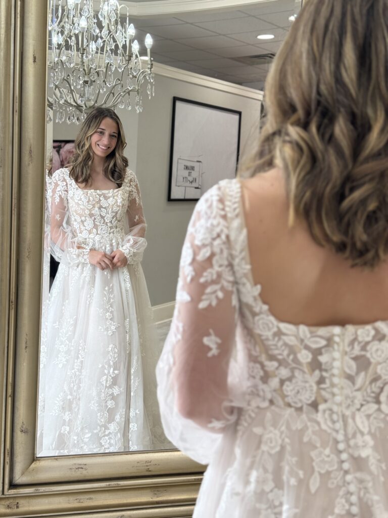 A bride smiles at her reflection while wearing a romantic lace wedding dress with long sheer sleeves and a square neckline at Amanda’s Touch.