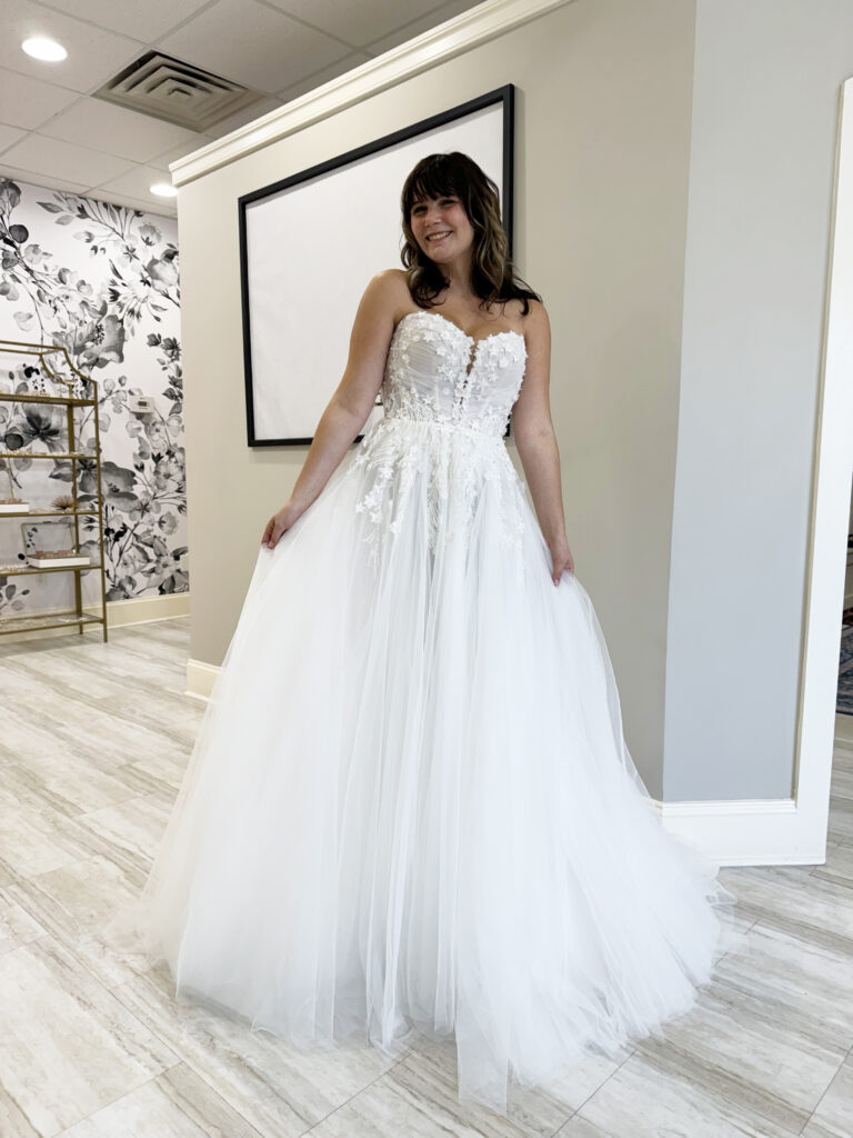A bride poses in a strapless ballgown wedding dress with a sweetheart neckline and a full tulle skirt, smiling inside Amanda’s Touch bridal shop.
