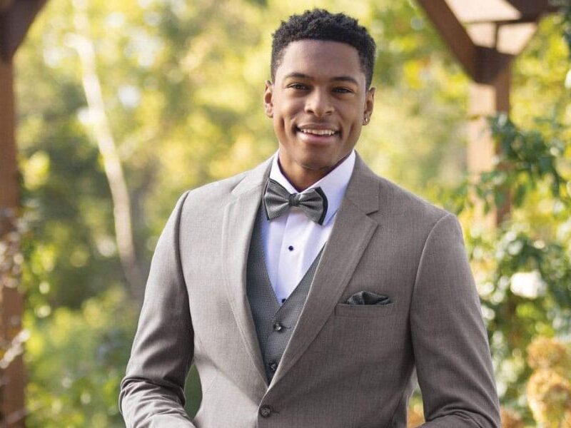 Close-up of a man in a tan slim fit tuxedo with a gray bow tie, standing in a garden with a relaxed and stylish expression.