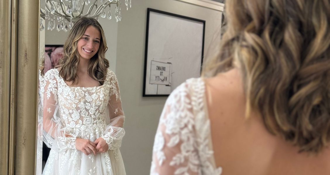A close-up of a bride smiling at her reflection while wearing a romantic long-sleeve lace wedding dress with floral embroidery at Amanda’s Touch.