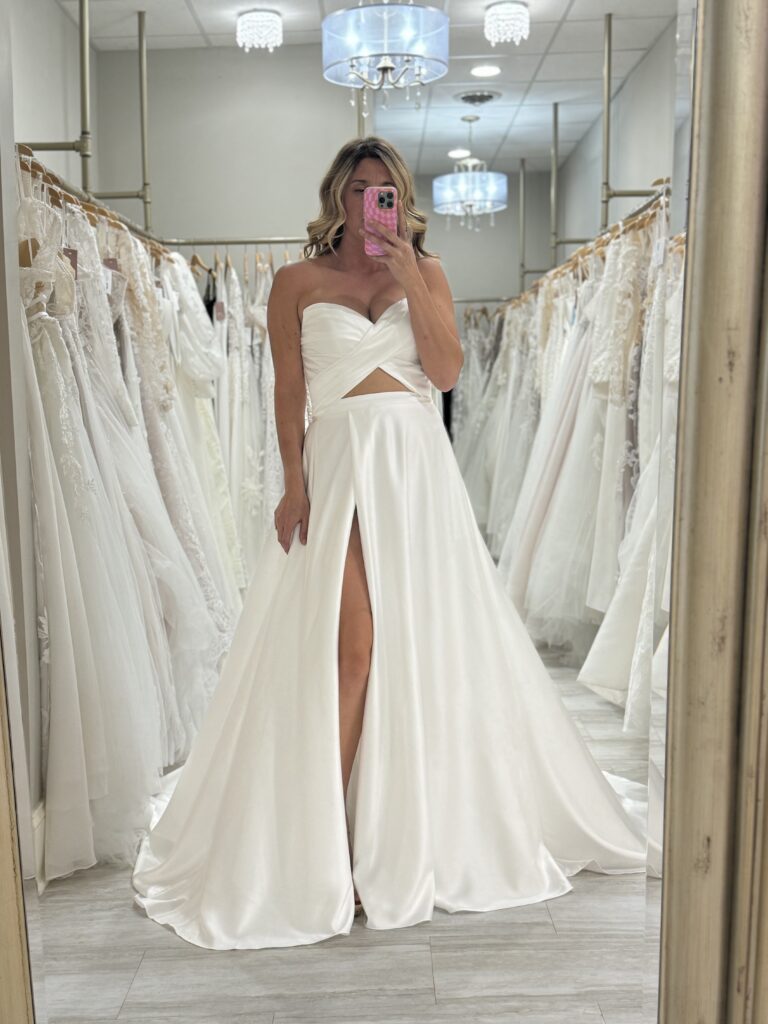 A bride takes a mirror selfie in a strapless satin wedding gown featuring a modern cut-out bodice and thigh-high slit inside Amanda’s Touch showroom.