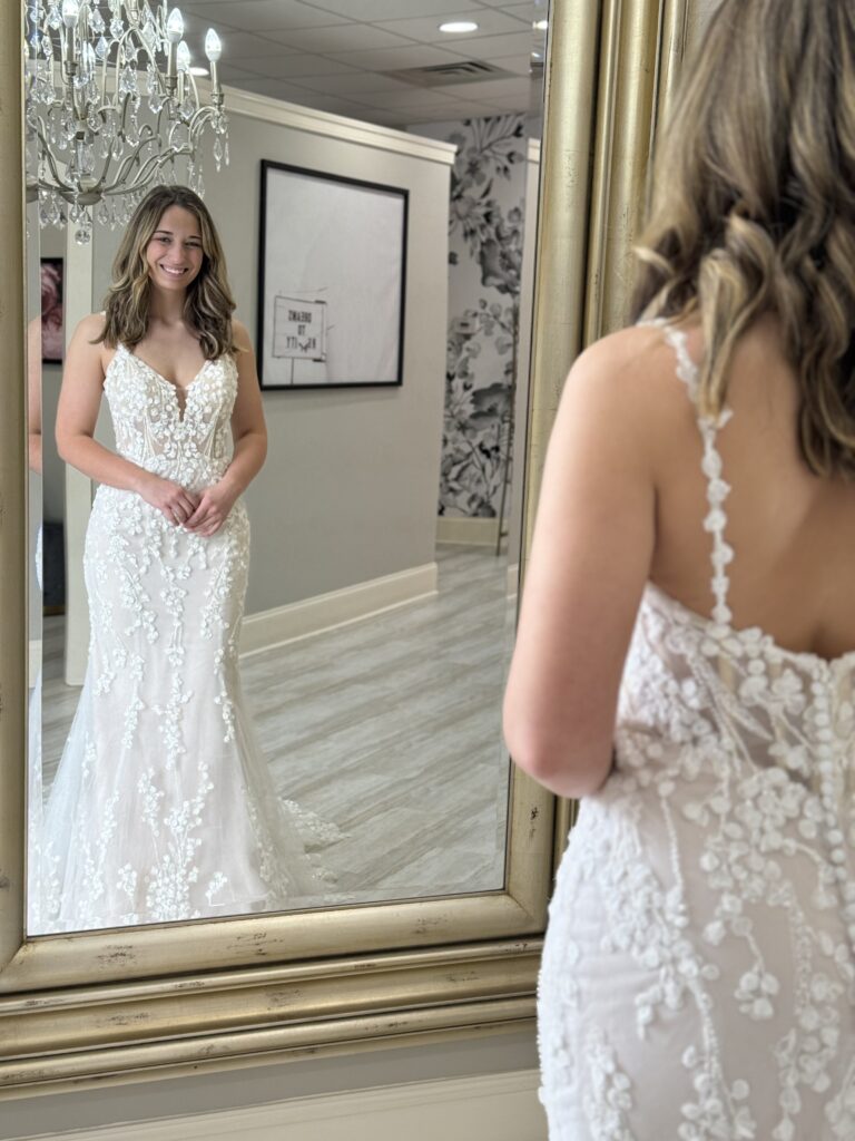 A bride stands in front of a mirror in a fitted lace wedding dress with thin straps and floral appliqué details, smiling at Amanda’s Touch bridal boutique.