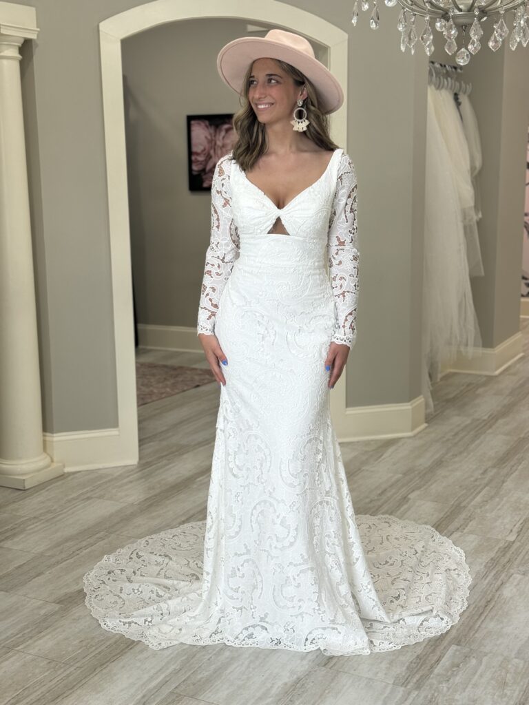 A bride models a boho-inspired lace wedding gown with long sleeves, a front cut-out, and a dramatic train, styled with a blush pink hat at Amanda’s Touch.