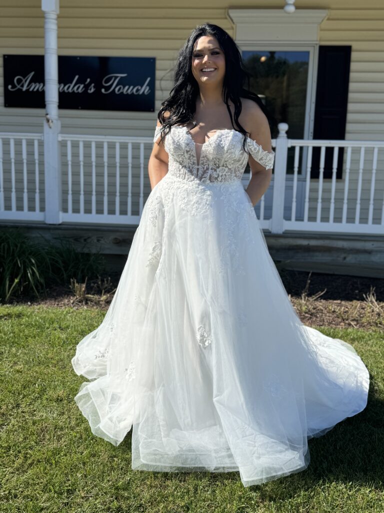 Bride standing outside Amanda’s Touch wearing an off-the-shoulder ballgown wedding dress with floral lace detailing and a sheer bodice.