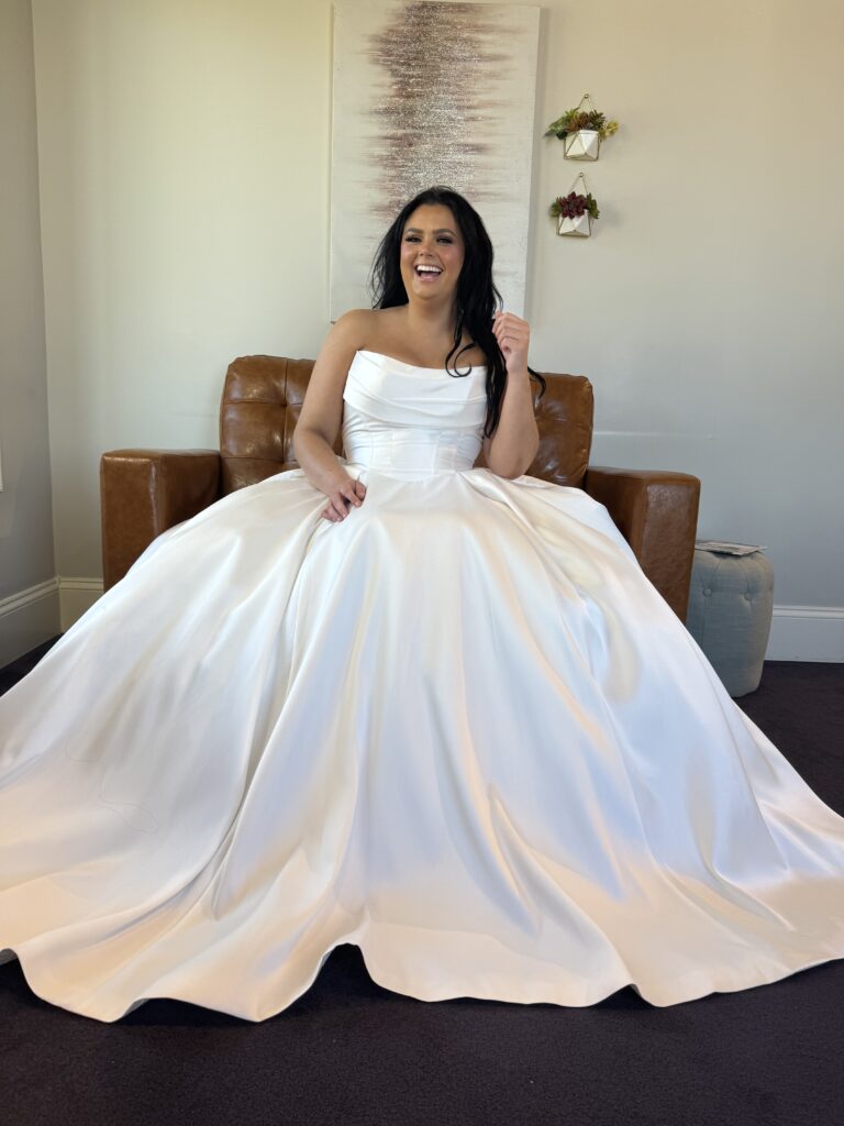 Bride sitting on a leather chair in a strapless satin ballgown wedding dress with a softly ruched bodice and full skirt at Amanda’s Touch.