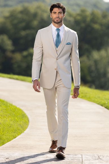 Groom walking down a curved outdoor path wearing a tan summer tuxedo with a teal tie and vest, hands relaxed at his sides.