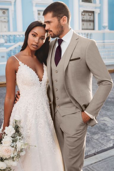 Groom in a sand-colored tuxedo with a burgundy tie and vest, standing with a bride in a lace gown in front of a pastel blue building.
