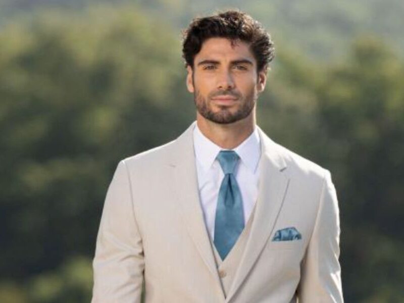 Groom wearing a tan summer wedding tuxedo with a teal tie and matching pocket square, standing outdoors with trees in the background.